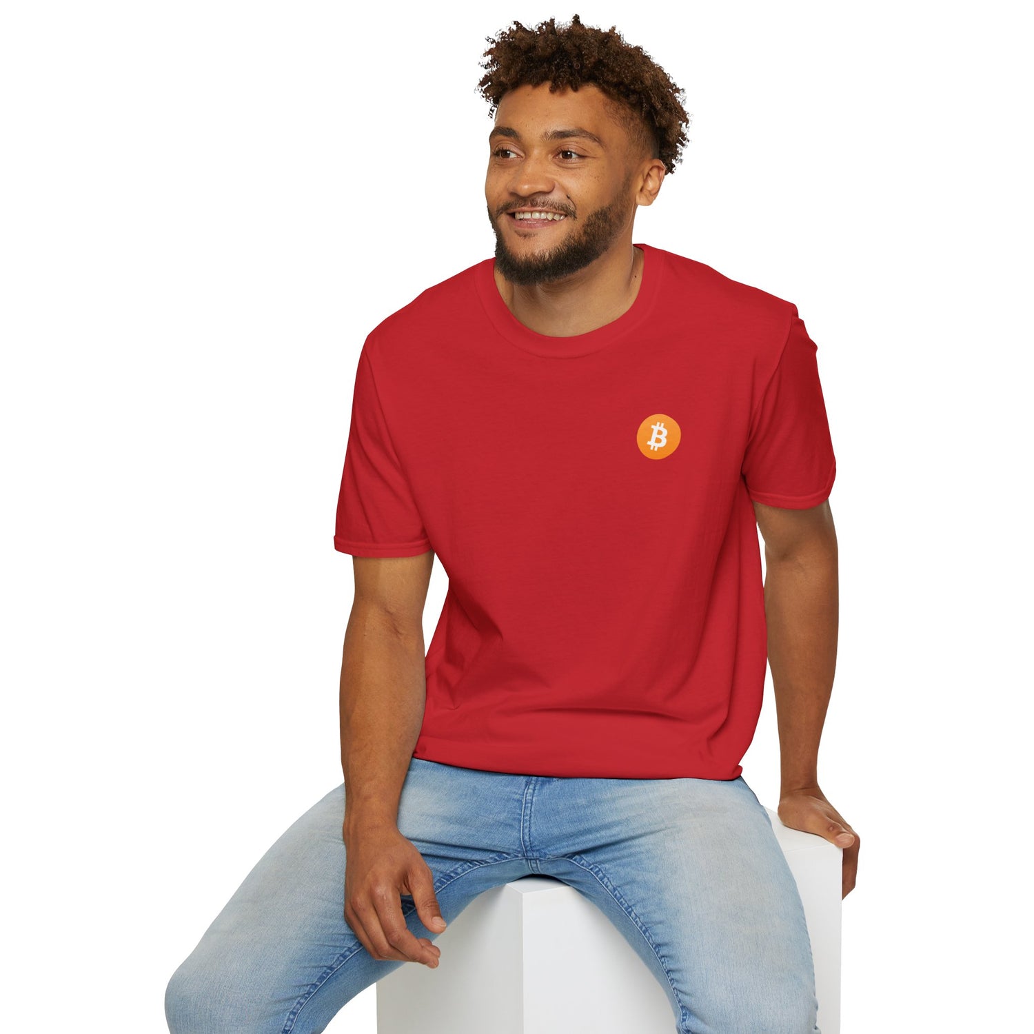 A guy sitting on a white cube smiling wearing a red round neck t-shirt with a small orange bitcoin logo on the left chest