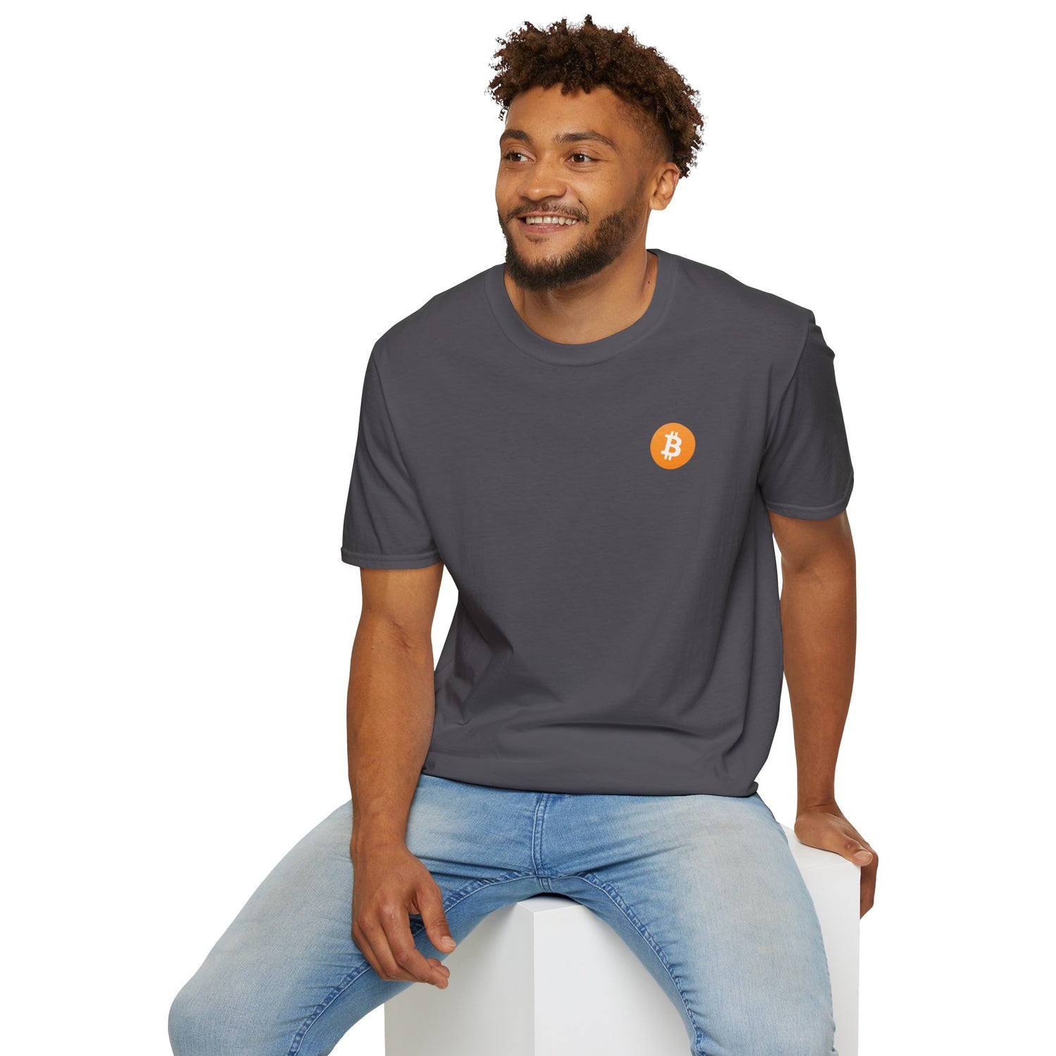 A guy sitting on a white cube smiling wearing a dark grey round neck t-shirt with a small orange bitcoin logo on the left chest