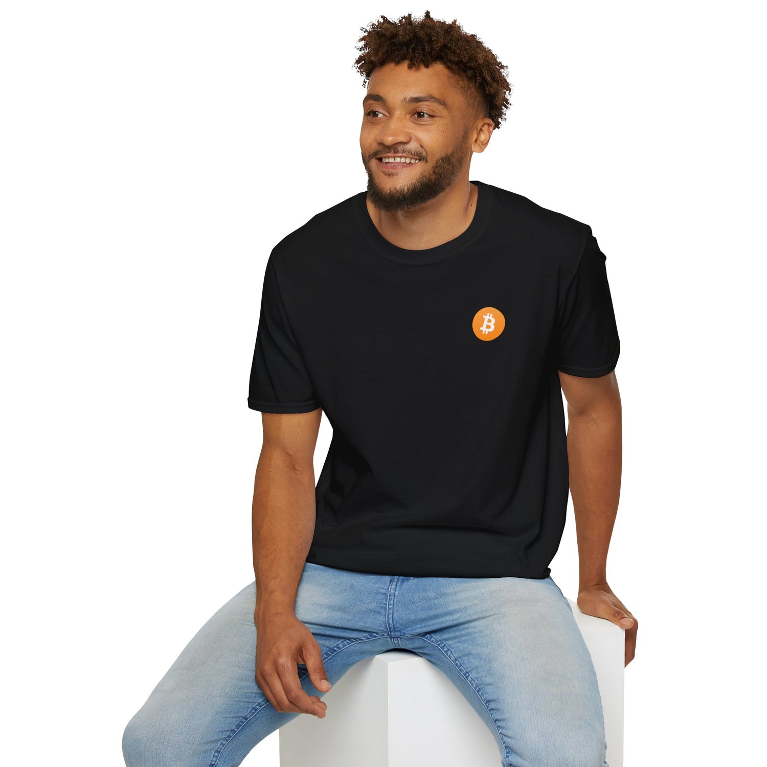 A guy sitting on a white cube smiling wearing a black round neck t-shirt with a small orange bitcoin logo on the left chest