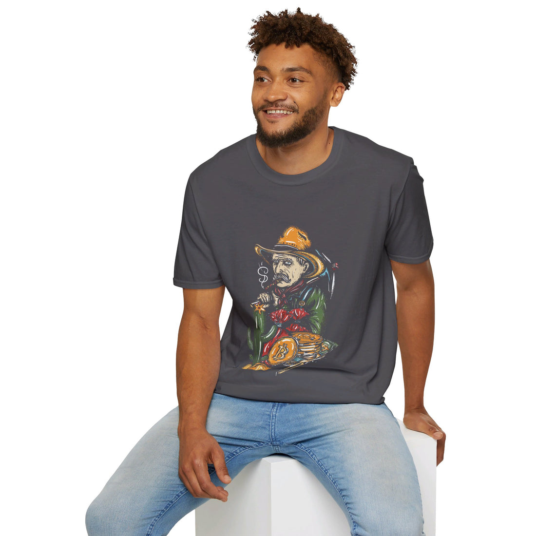 A man sits on a white block, smiling, wearing a dark grey T-shirt with a graphic of a skeleton in a cowboy outfit smoking a cigar and holding a bag of money, with a stack of bitcoin coins